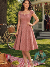 Pinup girl Woman in a red gingham dress standing outdoors with a picnic setup and bicycle in the background.