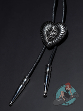 A black braided bolo tie with a textured, metallic silver heart-shaped centerpiece featuring a sunburst pattern and a small rose motif. The bolo tie has silver metal tips and opens into a locket