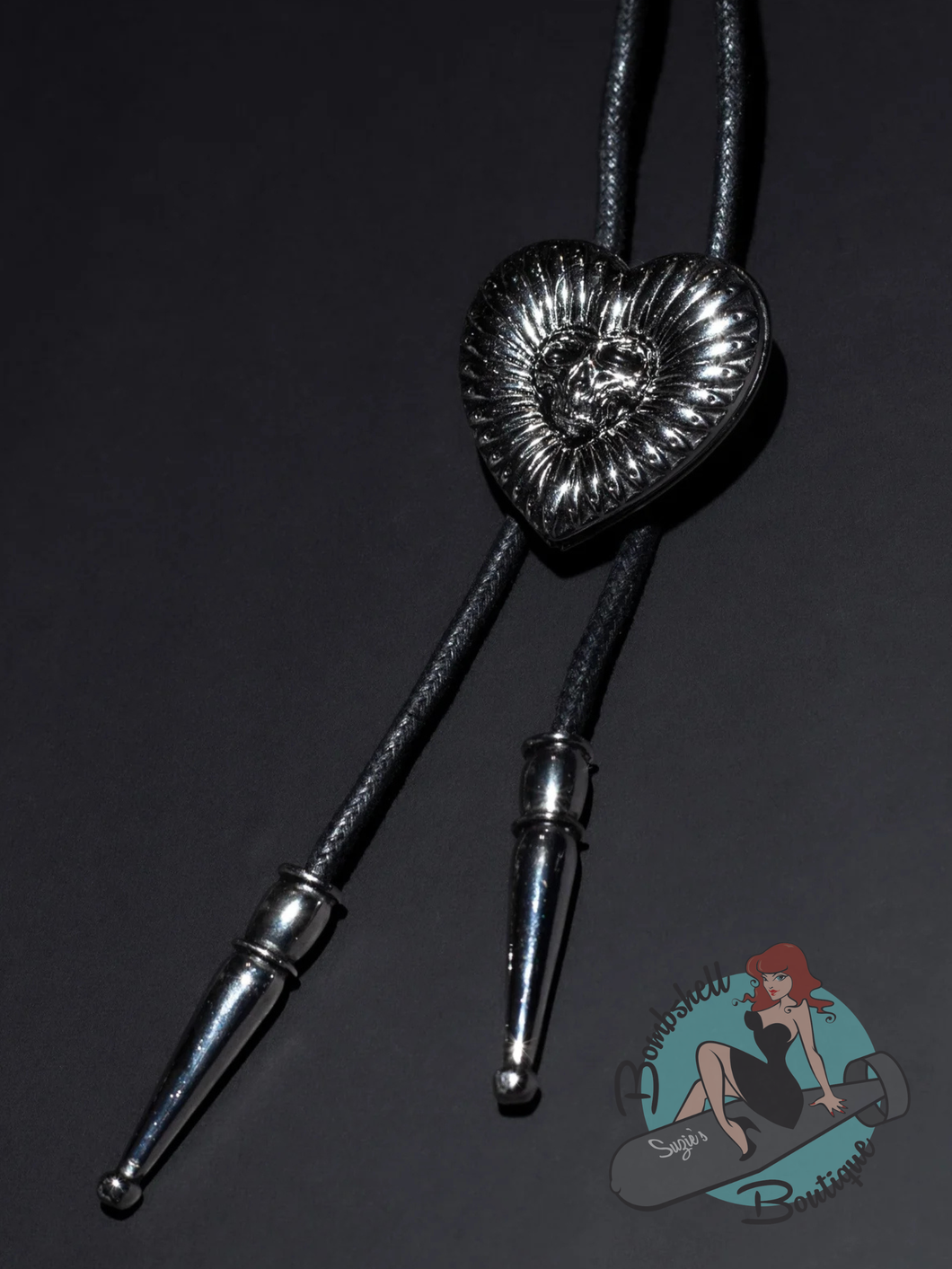 A black braided bolo tie with a textured, metallic silver heart-shaped centerpiece featuring a sunburst pattern and a small rose motif. The bolo tie has silver metal tips and opens into a locket
